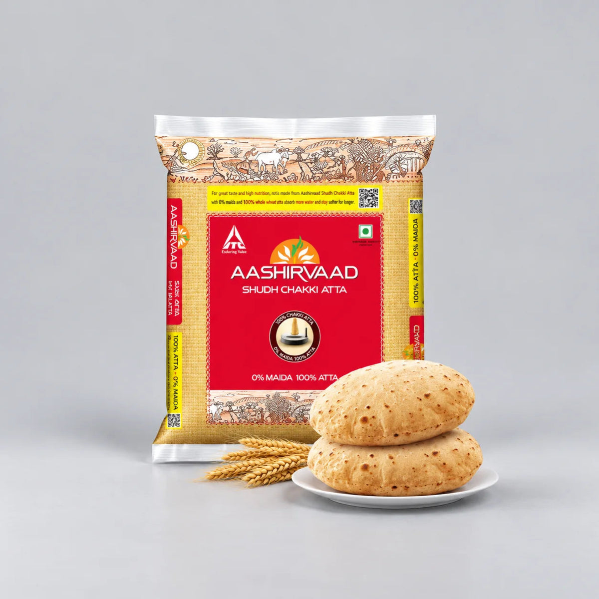 A bag of Aashirvaad Shujji Chaki Atta flour stands on a light gray studio background next to two round flatbreads stacked on a small white plate with a few wheat stalks. The packaging is primarily red and beige with yellow accents.