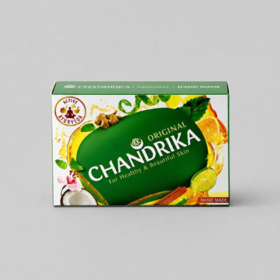 A rectangular box of Chandrika Original Hand Made soap, featuring a green and yellow design with illustrations of ingredients like ginger, citrus, and coconut, set against a neutral gray background.