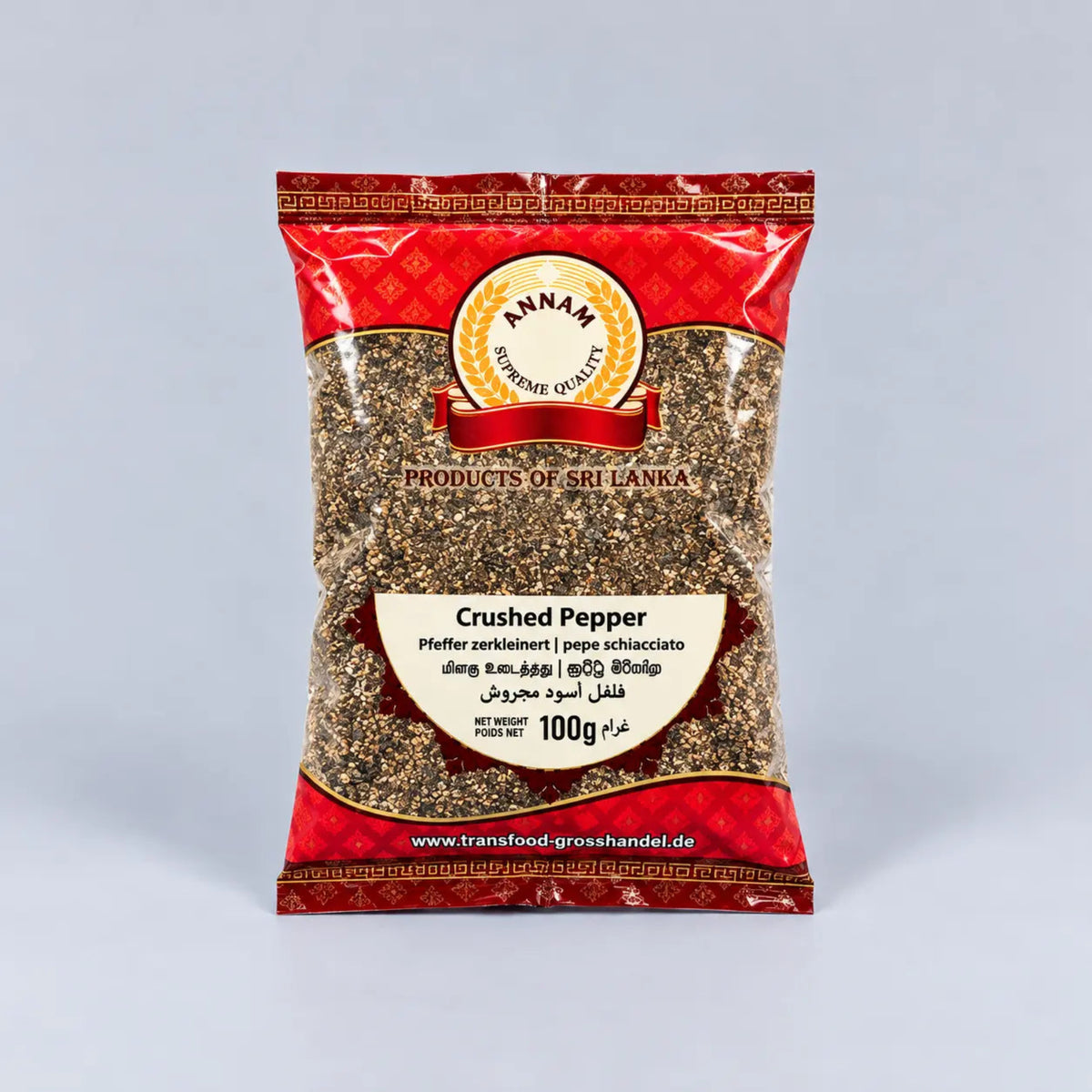 A 100g bag of Annam Crushed Pepper (pepper schiacciato) from Products of Sri Lanka, featuring a red and gold patterned packaging with a clear window showing the ground black pepper inside, set against a light gray background.
