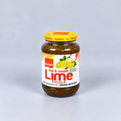 A glass jar of Eastern brand hot & sweet lime pickle, featuring a yellow lid and a white label with red and green text and lime illustrations, set against a neutral gray background.
