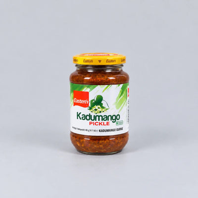 A glass jar of Eastern Kadumango Pickle (Kadumango Guri) with a yellow lid and a white label featuring green and red branding, set against a plain light gray background.