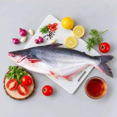 A whole raw fish laid on a white cutting board, surrounded by lemon slices, tomatoes, red onions, garlic, peppercorns, parsley, and a small glass of amber liquid on a light gray background.