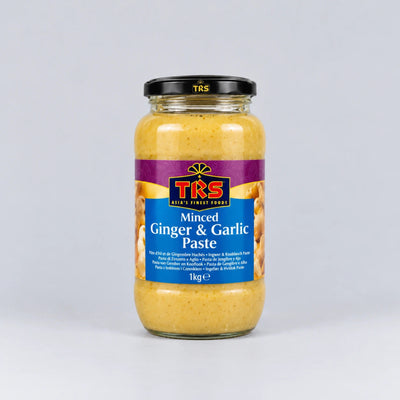 A 1kg glass jar of TRS Minced Ginger & Garlic Paste on a light gray background. The paste inside is visible through the clear glass, and the jar has a black lid with the TRS brand logo and a blue and purple label.