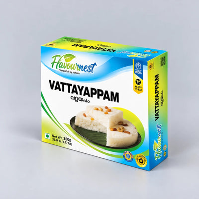 A box of Flavournest Vattayappam (Indian rice cake) shown at a three-quarter angle on a light gray background. The packaging is white with blue and green accents, featuring a photo of the white rice cake topped with brown seeds.
