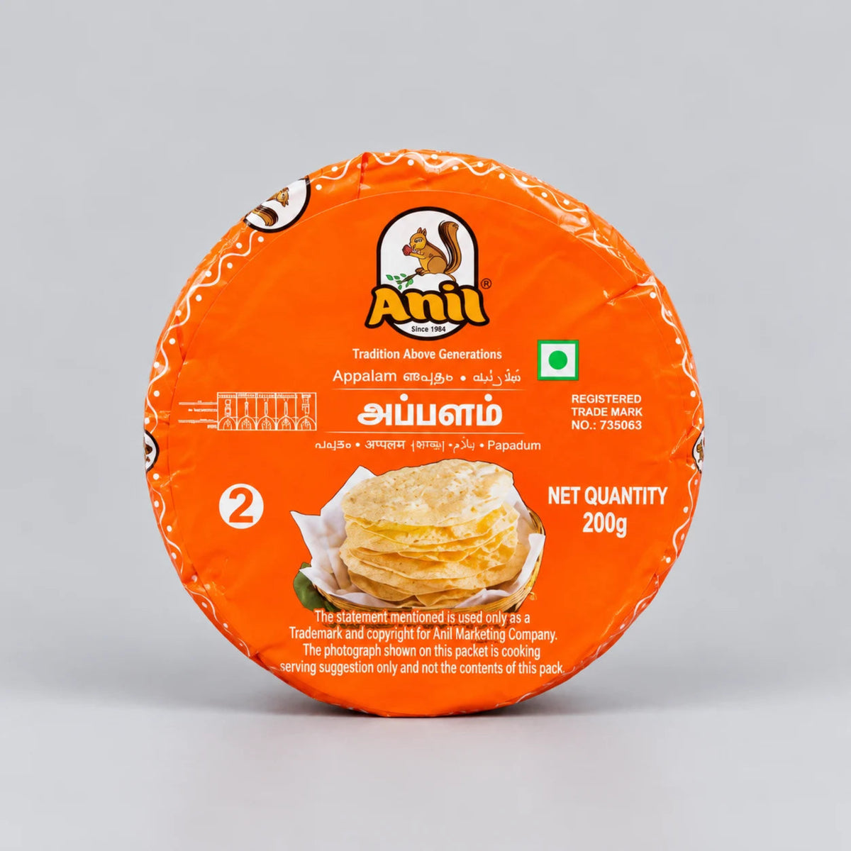 A round orange-wrapped food product on a light gray studio background. The packaging prominently shows the brand name 'Anil' and the product name 'Appalam' with a photo of layered savory pancakes on the front.