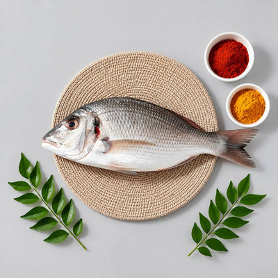 A whole silver fish laid on a round woven beige placemat, with two small white bowls of red and yellow spices at the upper right and green leafy stems at the lower corners, all on a light gray background.